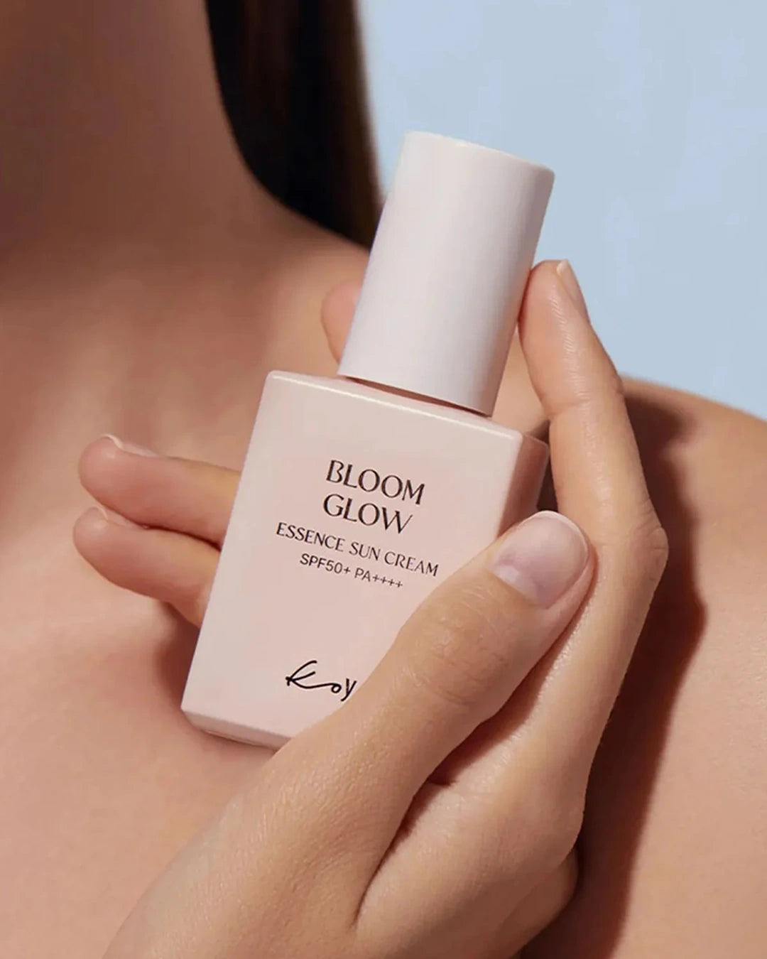 A close-up of a person holding a pale pink bottle of Kiehl's Bloom Glow Essence Sun Cream SPF 50+ against a soft light blue background.