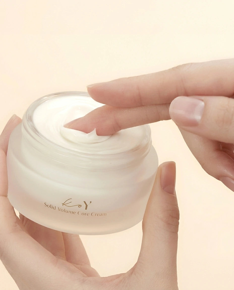 A hand holding an open jar of solid volume care cream, with another hand touching the cream inside, set against a soft beige background.