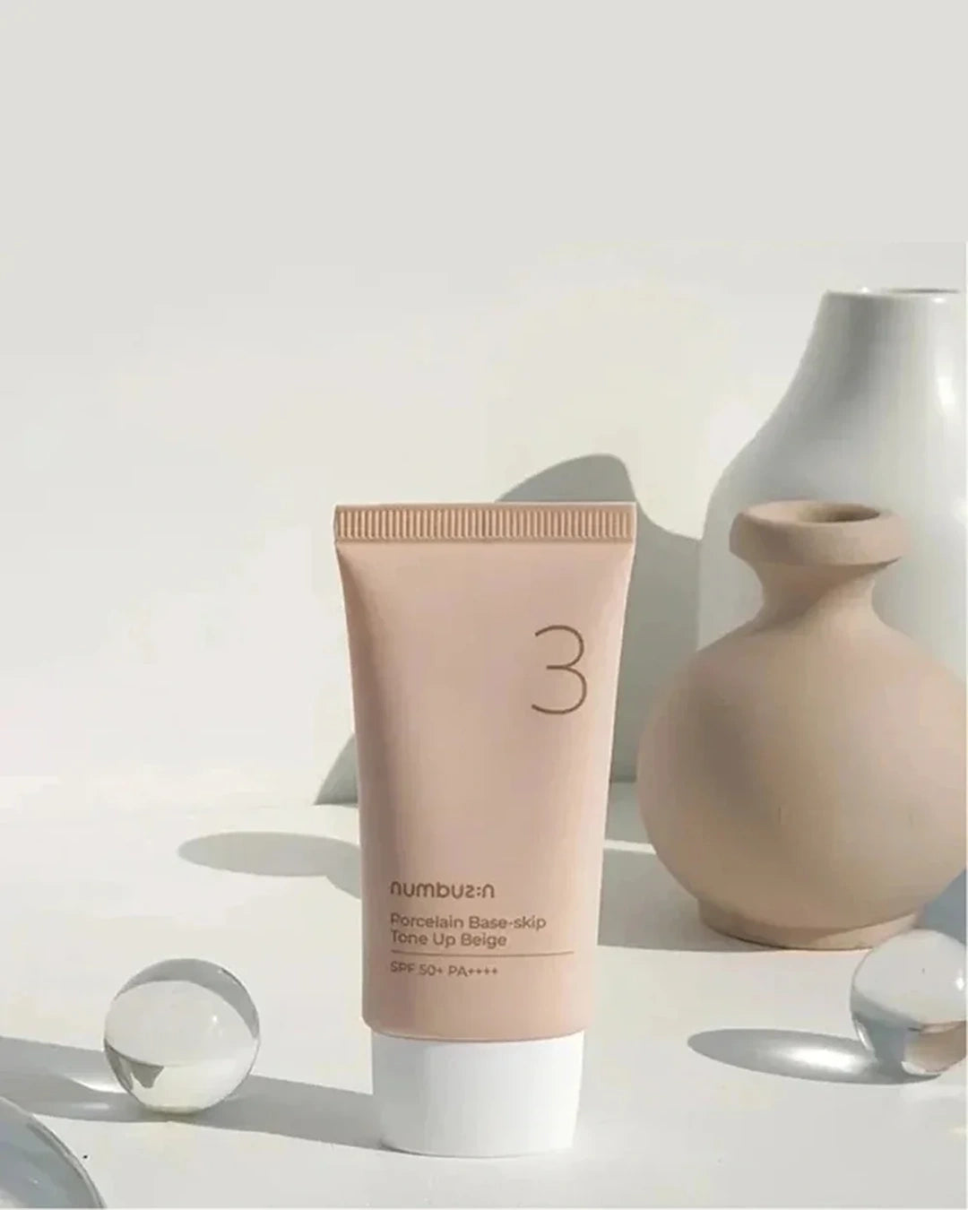 A squeeze tube of numbuen Porcelain Base-skip Tinted Lip Balm shade 3, standing upright on a white surface with decorative ceramic vases and glass spheres in the background.