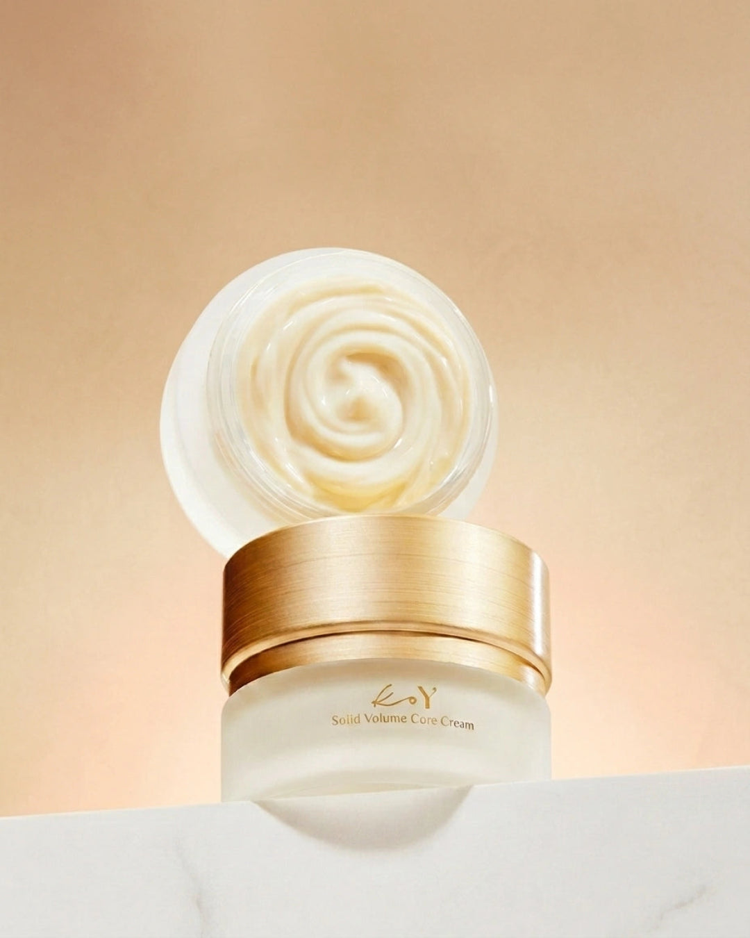 A jar of KAY Solid Volume Care Cream with its lid off, showing a swirl of creamy off-white product inside. The jar has a metallic gold lid and a frosted white base, set against a warm beige background.