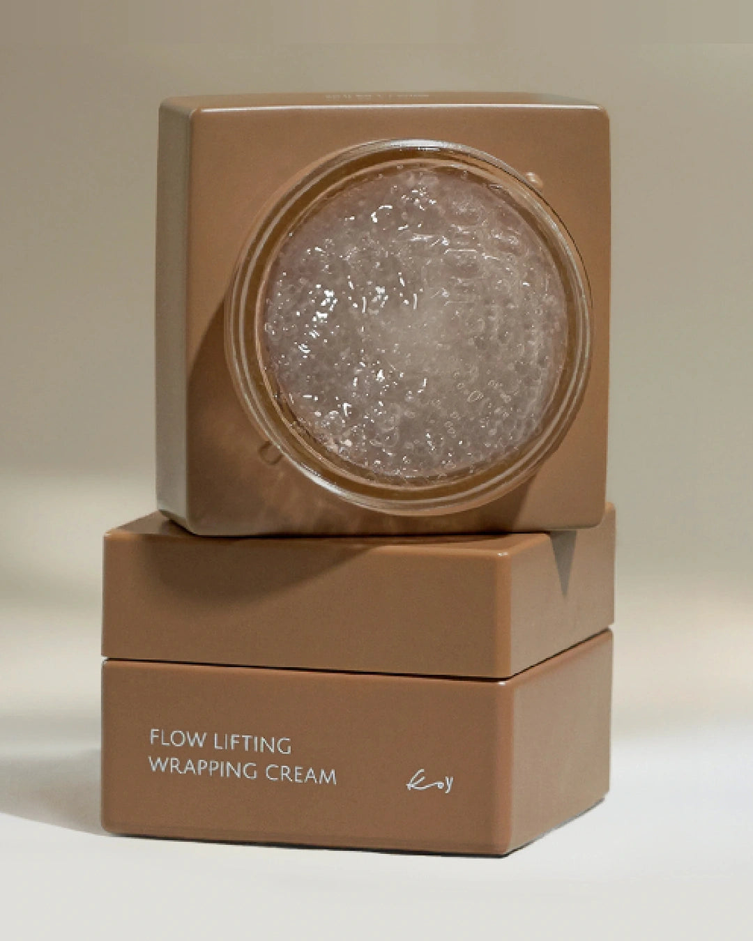 A stacked cosmetic jar set labeled 'FLOW LIFTING WRAPPING CREAM' in a warm taupe-brown color, with the top jar open to reveal a clear gel-like cream inside, on a neutral beige background.
