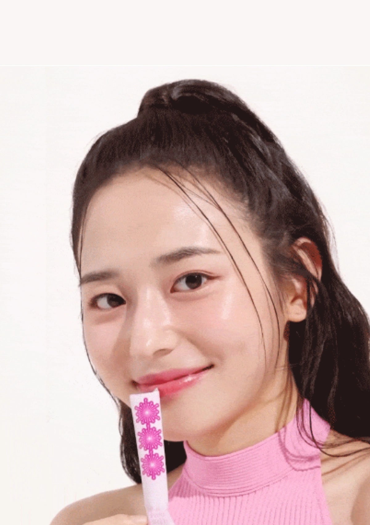 A close-up portrait of a woman with dark hair in a high ponytail, wearing a pink mock-neck top and holding a pink and white flower-patterned stick against her teeth, set against a plain white background.