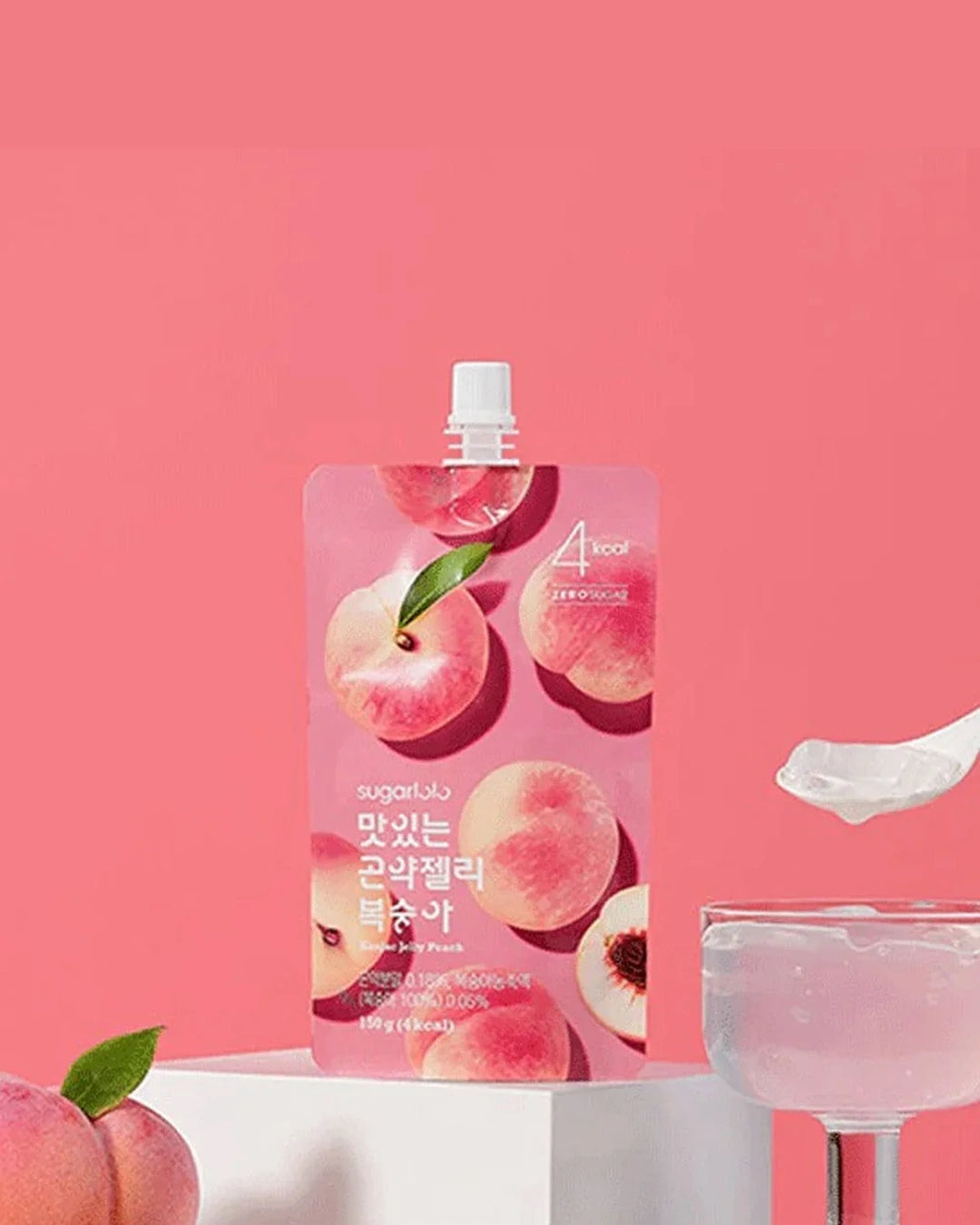 A pink stand-up pouch of a peach-flavored cosmetic or skincare product with peach illustrations and Korean text, displayed on a white pedestal against a pink background. A spoonful of pale pink product and a glass of pale pink liquid are shown beside it.