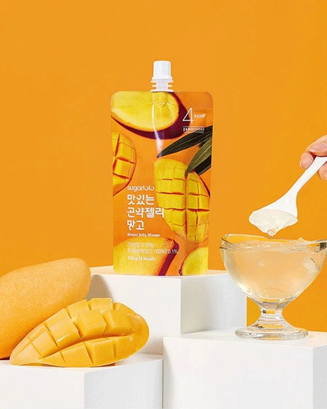 A stand-up pouch of Tangerine flavor drink mix by Four Senses stands on white display blocks next to a glass bowl with a spoonful of the pale yellow liquid, with fresh mangoes in the foreground against a solid orange background.