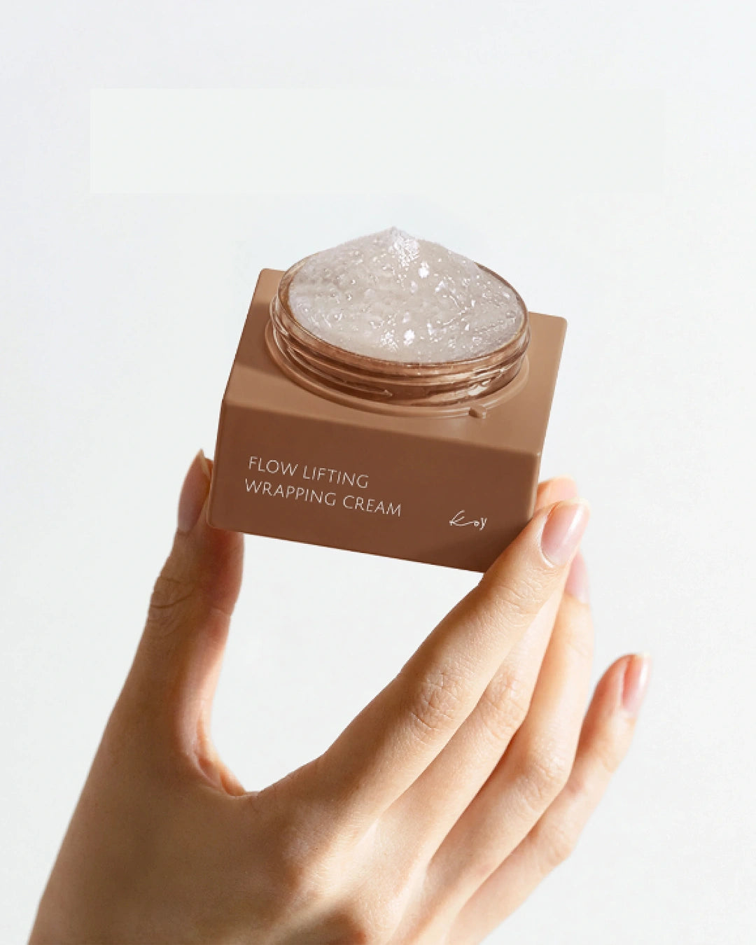 A hand holding a small square brown jar of Flow Lifting Wrapping Cream against a clean white background.