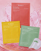 Three Biodance Real Deep Mask sachets in coral pink, yellow, and mint green are arranged on a pink water-splash background, with a translucent white sheet mask placed in front.