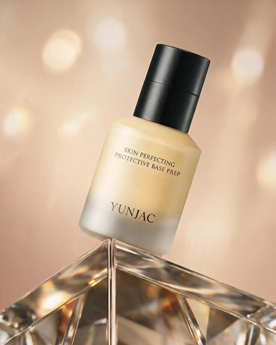 YUNJAC Skin Perfecting Protective Base Prep in a frosted glass bottle with a black cap, displayed on a faceted crystal pedestal against a warm, softly blurred bokeh background.