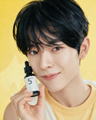 A close-up of a young man with dark hair holding a small dropper bottle labeled '5+' against a bright yellow background.