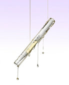 A clear glass cosmetic tube containing a pale yellow liquid, suspended diagonally with dripping glass and liquid details against a soft lavender-to-white gradient background.