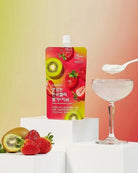 A stand-up pouch of strawberry and kiwi flavored drink mix stands on white pedestals beside a coupe glass filled with the pale pink drink and a spoon, with whole and sliced strawberries and a kiwi placed in front. The pouch has a bright pink-to-yellow gradient with fruit illustrations and Korean text.