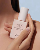 A close-up of a person holding a pale pink bottle of Kiehl's Bloom Glow Essence Sun Cream SPF 50+ against a soft light blue background.