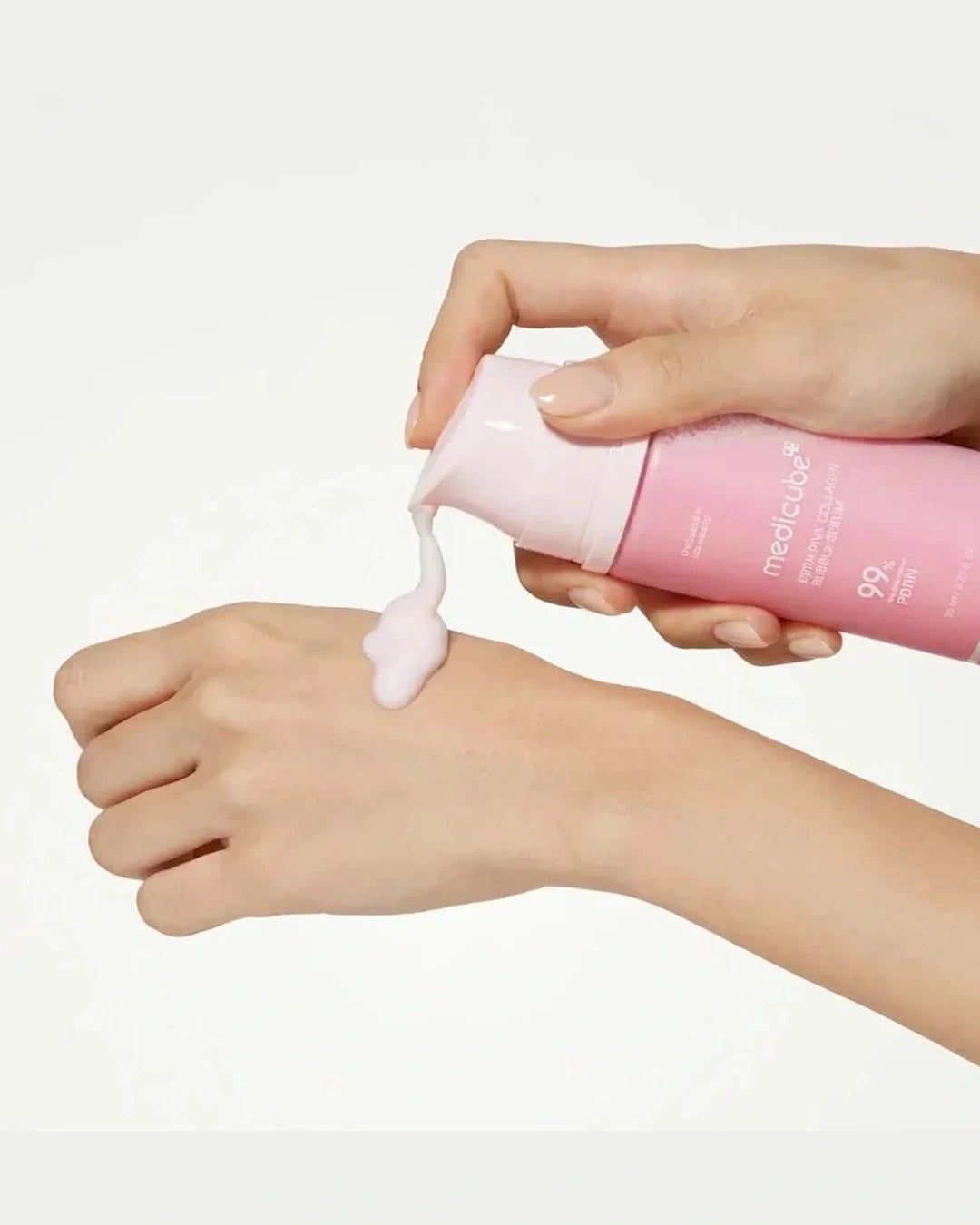 A close-up of a person's hands dispensing a pale pink cream from a pink tube onto the back of another hand, against a clean white background.