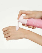 A close-up of a person's hands dispensing a pale pink cream from a pink tube onto the back of another hand, against a clean white background.