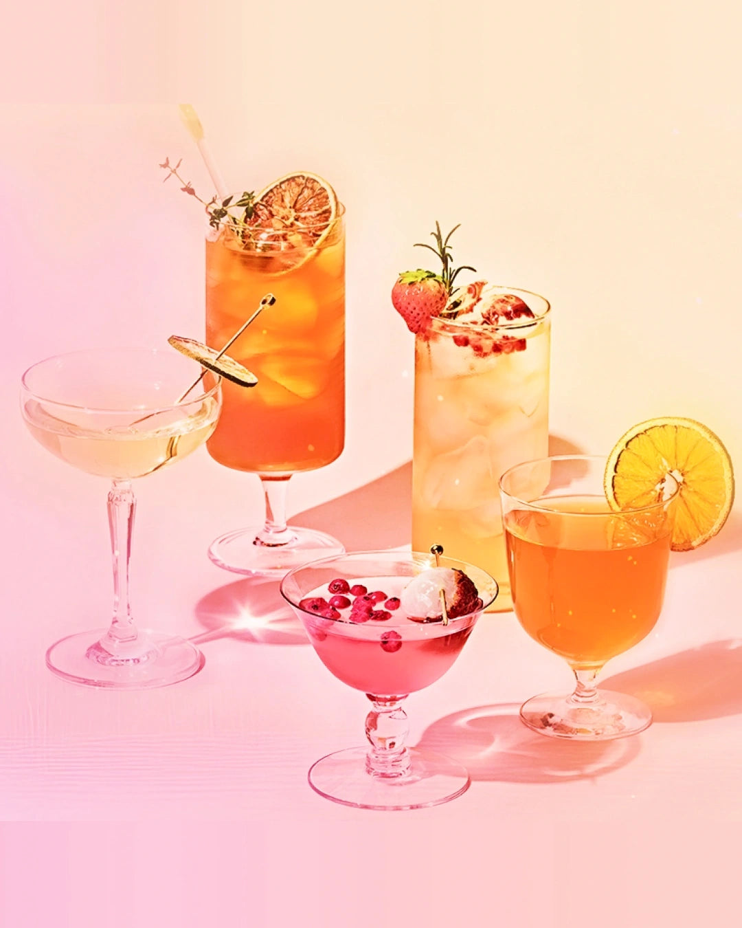 Five colorful cocktails in various glassware, including a coupe, highball, and martini glasses, arranged on a pink and peach gradient background with soft shadows.