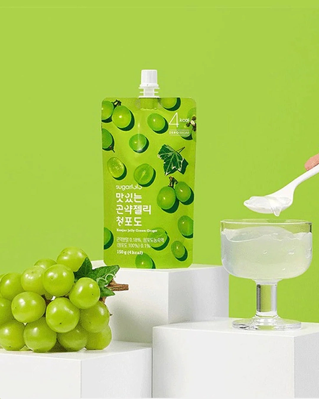 A green squeeze pouch of grape-flavored drink mix stands on white pedestals against a bright green background, with a bunch of green grapes, a glass of pale green liquid, and a hand holding a spoon with white powder.