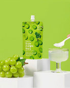 A green squeeze pouch of grape-flavored drink mix stands on white pedestals against a bright green background, with a bunch of green grapes, a glass of pale green liquid, and a hand holding a spoon with white powder.