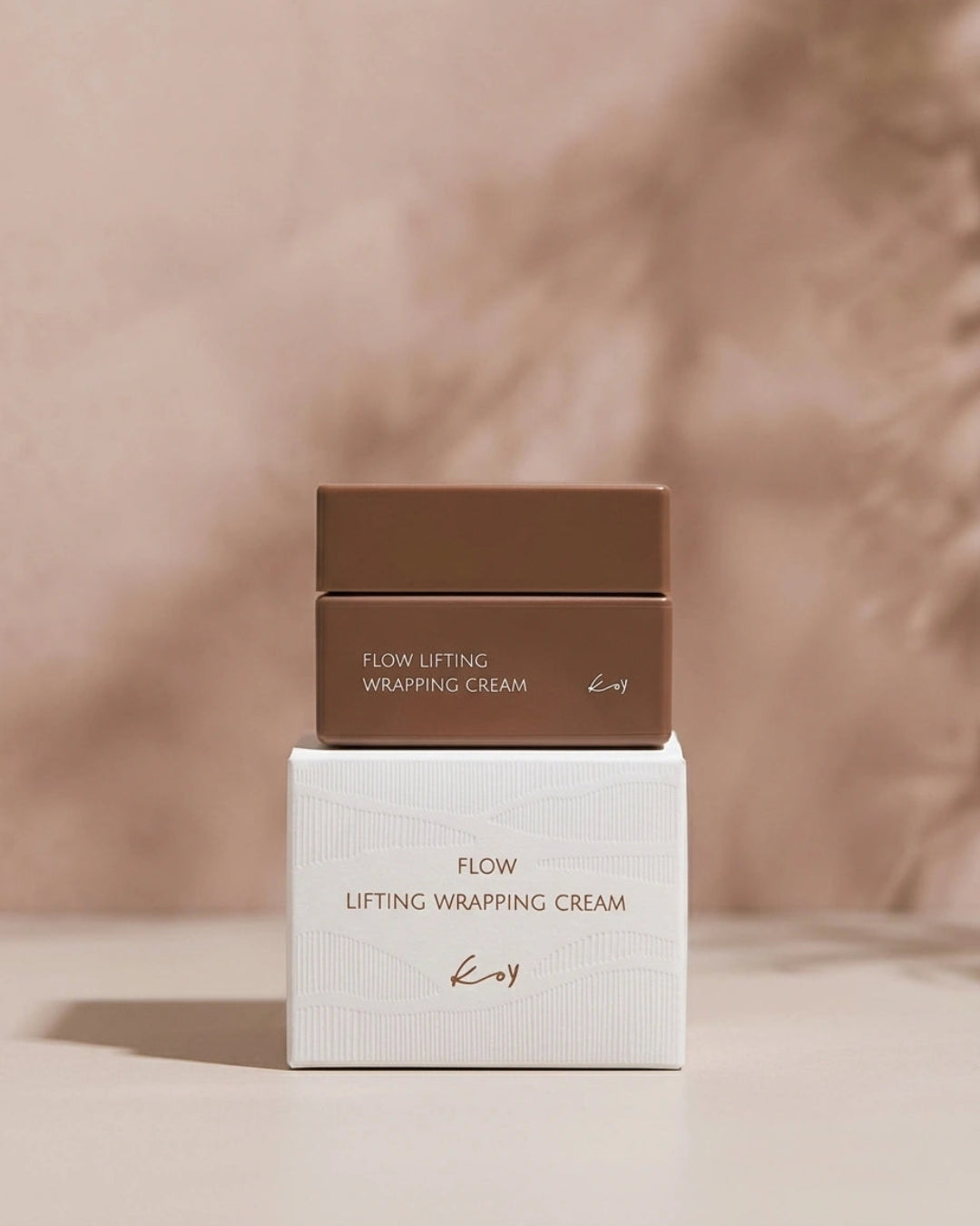 A cosmetic cream jar in a medium brown shade sits on top of its white retail box, both printed with the text 'FLOW LIFTING WRAPPING CREAM' and a small 'kv' logo. The set is photographed on a neutral beige surface against a soft, textured beige background.