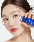 A close-up of a woman applying cream from a blue Medlabce skincare pump bottle to her cheek against a clean white background.
