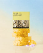 A stack of yellow-orange gummy candies with a pale yellow NE:AR brand card standing behind them, plus three small lavender round gummies in front, set against a soft blue-to-beige gradient background.