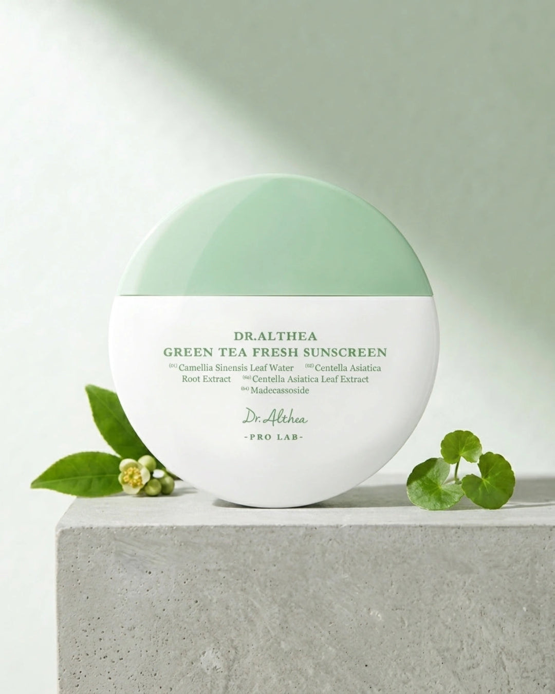 Dr. Althaea Green Tea Fresh Sunscreen in a round white jar with a mint green lid, styled on a light gray stone block with green leaves and small flowers against a soft light green background.