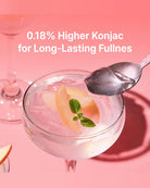 A pink-toned promotional shot of a stemmed glass filled with a pale pink beverage, garnished with peach slices and a mint leaf, with a spoon resting on the rim. White text at the top reads, '0.