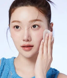 A close-up of a woman with her hair pulled back, wearing a bright blue knit top and using a white makeup sponge on her cheek against a light blue background.