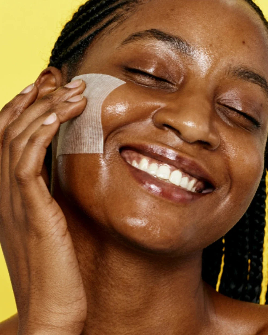 A close-up of a woman with braided hair smiling while applying a translucent adhesive patch to her cheek against a bright yellow background.