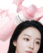 A close-up beauty shot of a woman with pink skincare cream dripping from her cheek, with a pink bottle of Medicube Hydro Cell-Massage cream floating above her head against a white background with pink liquid splashes.