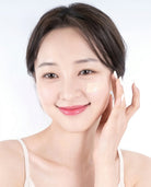 A close-up of a woman with dark brown hair applying or showing a light beige cream product on her cheek against a clean white background.