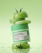 A green tomato-shaped deep bubble mask bottle with sliced tomato props, photographed on a light green background with water droplets and a reflective surface.