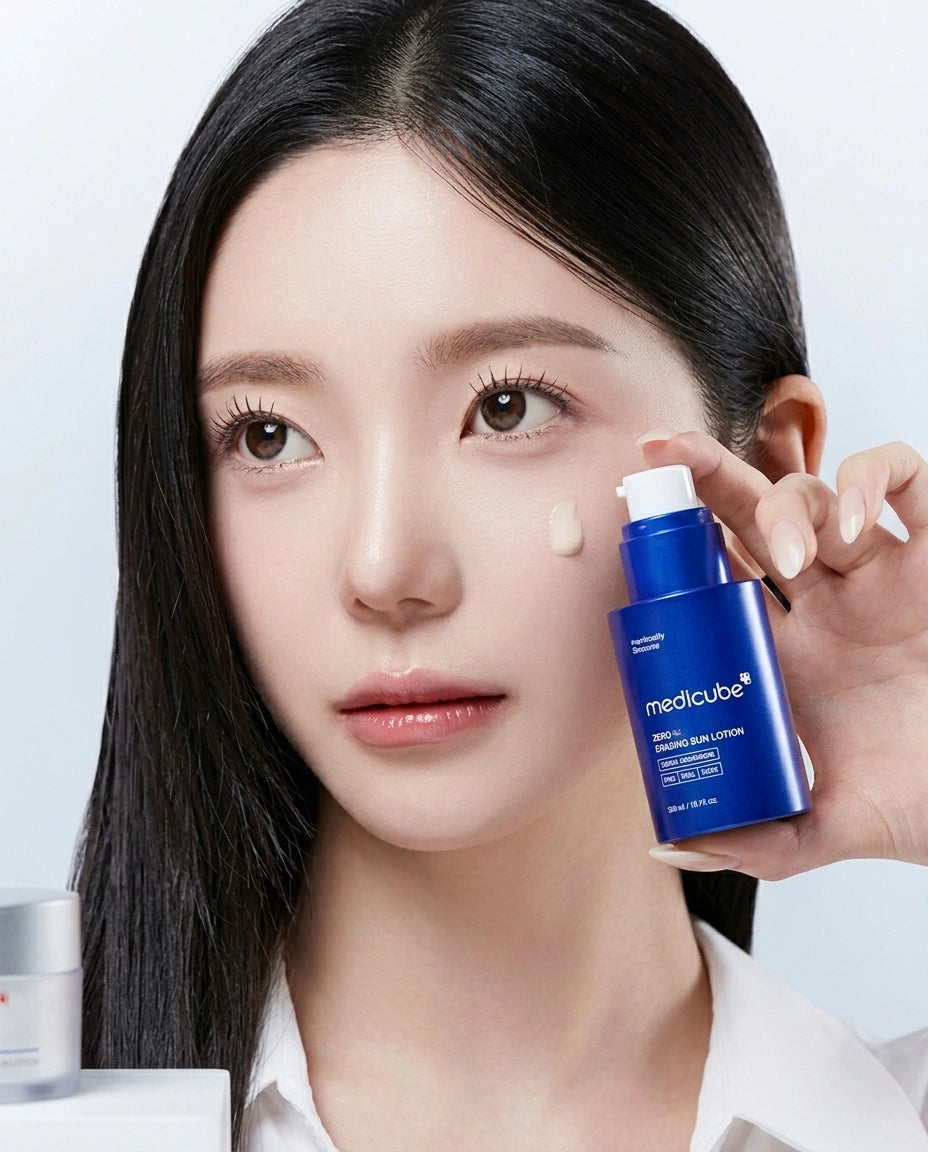 A close-up of a woman with long black hair holding a blue Medicaube skincare pump bottle next to a small dollop of cream on her cheek, against a clean light gray background.