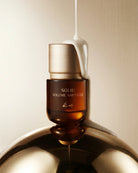 A cosmetic ampoule bottle labeled 'SOLID VOLUME AMPOULE' in amber glass with a metallic gold cap, shown with a white cream-like substance dripping into and around it on a warm beige background.