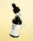 A small dark glass dropper bottle with a black rubber-and-plastic dropper cap, shown floating against a pale yellow background. The bottle has a white label with large black '5+' text and smaller text reading 'Citruslype Vitamin Concentrate' and 'Aumblon'.