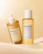 Two SKIN1004 Herbal Essence Centella skincare bottles on a soft beige fabric surface with a warm beige background. One bottle is a clear dropper bottle containing golden-yellow serum, and the other is a cylindrical toning tower bottle filled with pale yellow liquid.