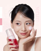 A woman with dark hair pulled back holds a pink and white skincare serum bottle while applying a white cream to her cheek against a soft pink background.