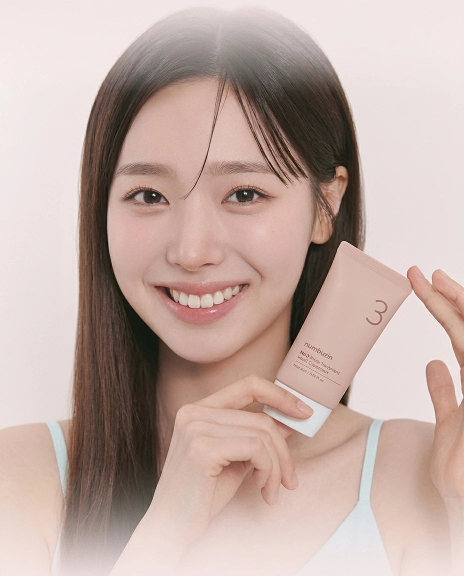 A smiling woman with long dark hair holds a light pink cosmetic tube near her face against a soft pale pink background.