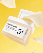 A jar of numbuzin 5+ Glutathione Vitamin Concentrated Toner Pads with a white lid, shown open on a bright yellow background with floating cream-colored pads.