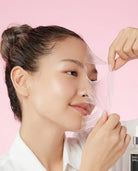 A woman with her hair in a bun applies a transparent peel-off mask to her face, pulling it away with her fingers against a solid pink background. A white skincare bottle labeled 'SKIN SERUM' is partially visible in the bottom right corner.