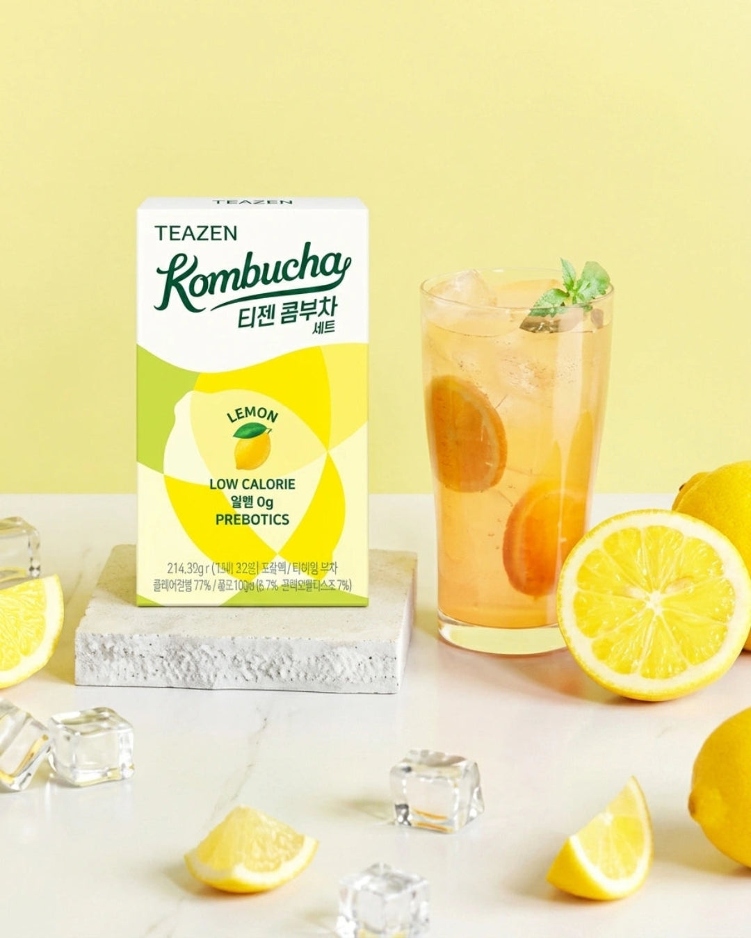 A box of TEAZEN Kombucha Lemon probiotics sits on a stone slab next to a glass of orange-tinged iced drink, surrounded by fresh lemons and ice cubes on a light surface against a pale yellow background.