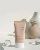 A squeeze tube of numbuen Porcelain Base-skip Tinted Lip Balm shade 3, standing upright on a white surface with decorative ceramic vases and glass spheres in the background.
