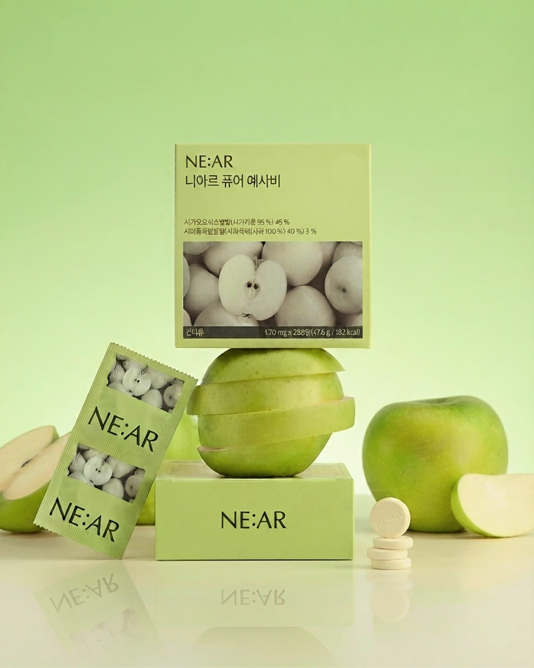 NE:AR pear skincare packaging arranged on a pale green reflective surface, with a rectangular box, a smaller angled box, a green pear, a sliced pear, and two white capsules. The product branding NE:AR is clearly visible on the packaging.