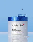 A white cosmetic jar with a metallic blue lid labeled medicube ZERO PORE PAD 2.0, shown against a light blue background with water droplets and a small splash above the container.