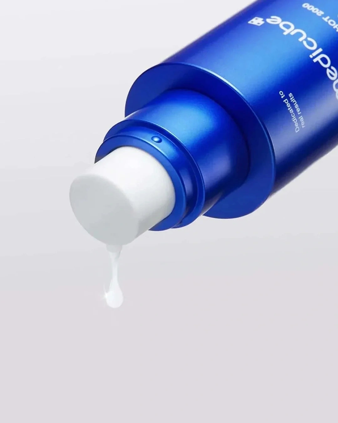 A close-up of a blue cosmetic bottle with a white pump dispensing a small drop of white cream against a light gray background.