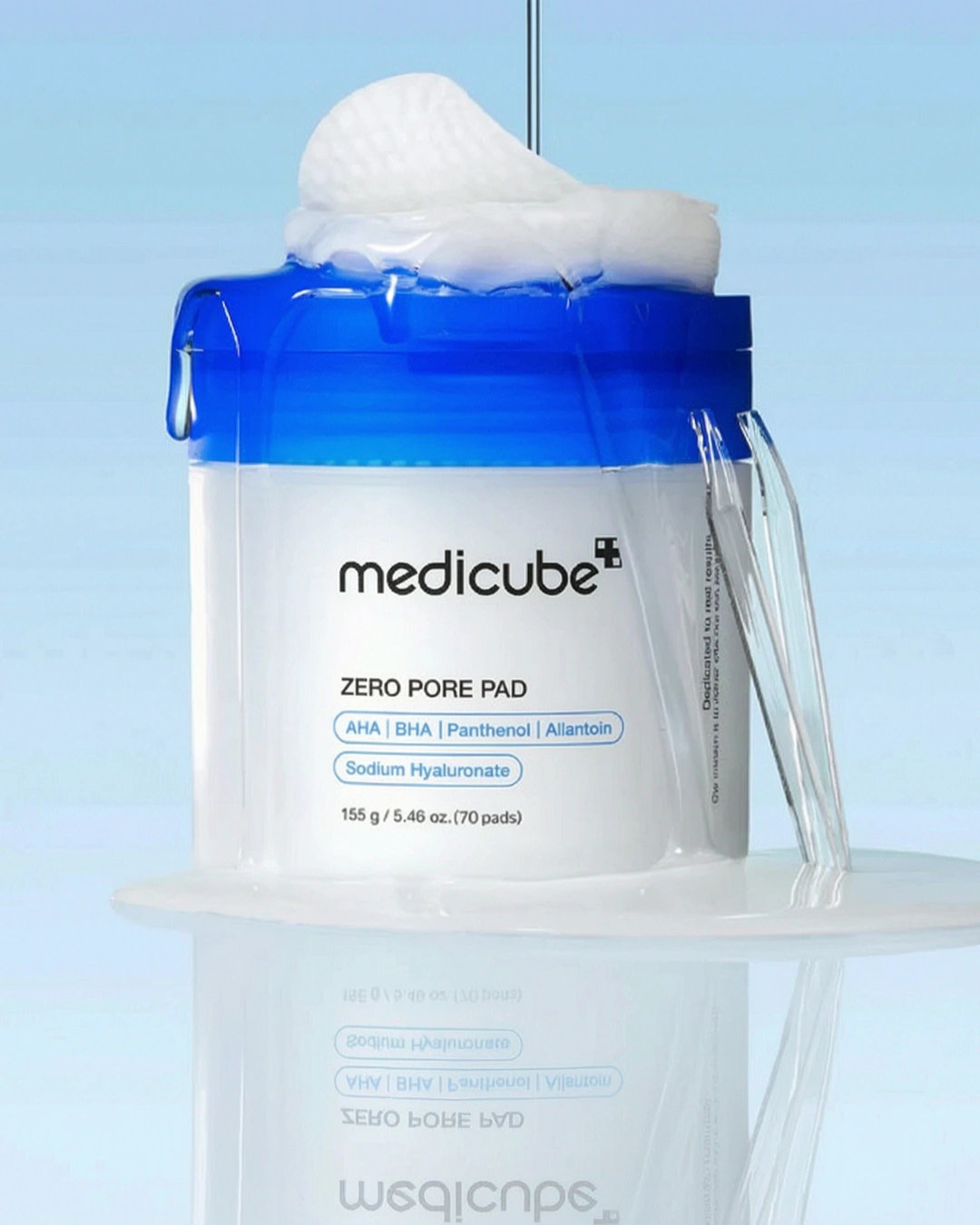 A white and blue jar of medicube+ Zero Pore Pad shown with a pile of white pads on top and a clear applicator, set against a light blue gradient background with a glossy reflection.