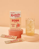 TEAZEN Kombucha Yogurt box in peach and white packaging, shown with two sachets and a glass of iced tea on pastel display pedestals against a warm beige background.