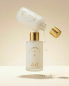 A frosted glass skincare bottle with a metallic gold cap, shown with a second tilted bottle pouring clear liquid into it. The bottles have gold lettering and are set against a warm beige background with a few floating water droplets.