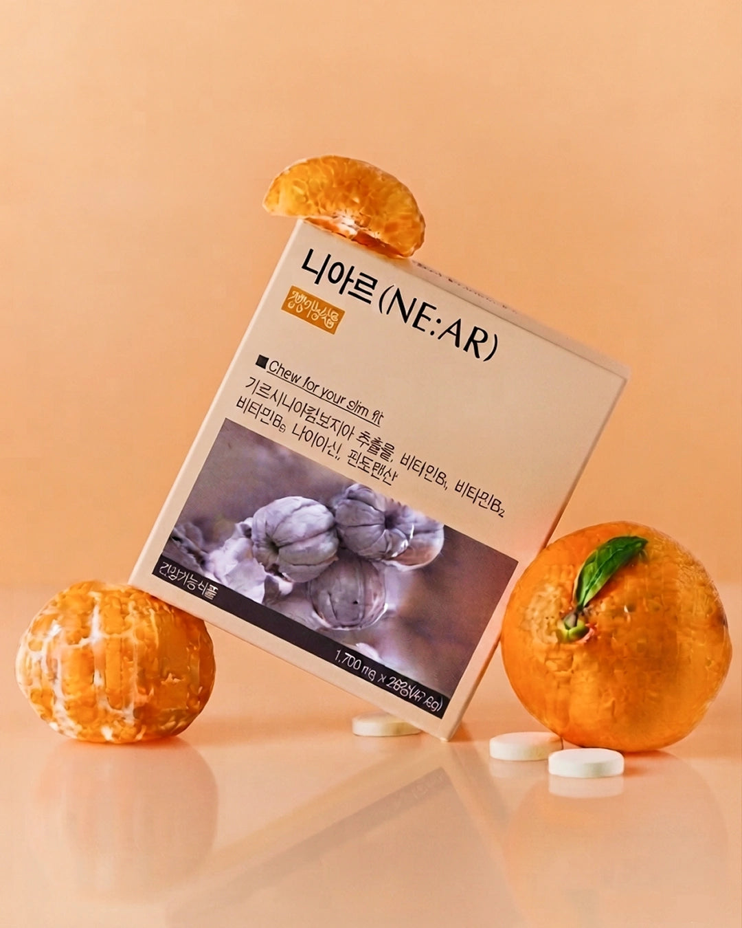 A box of NE:AR Nairine capsules styled with fresh orange slices and whole oranges on a warm peach background. The box features Korean text and a photo of garlic bulbs, with several white capsules scattered in front.