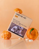 A box of NE:AR Nairine capsules styled with fresh orange slices and whole oranges on a warm peach background. The box features Korean text and a photo of garlic bulbs, with several white capsules scattered in front.
