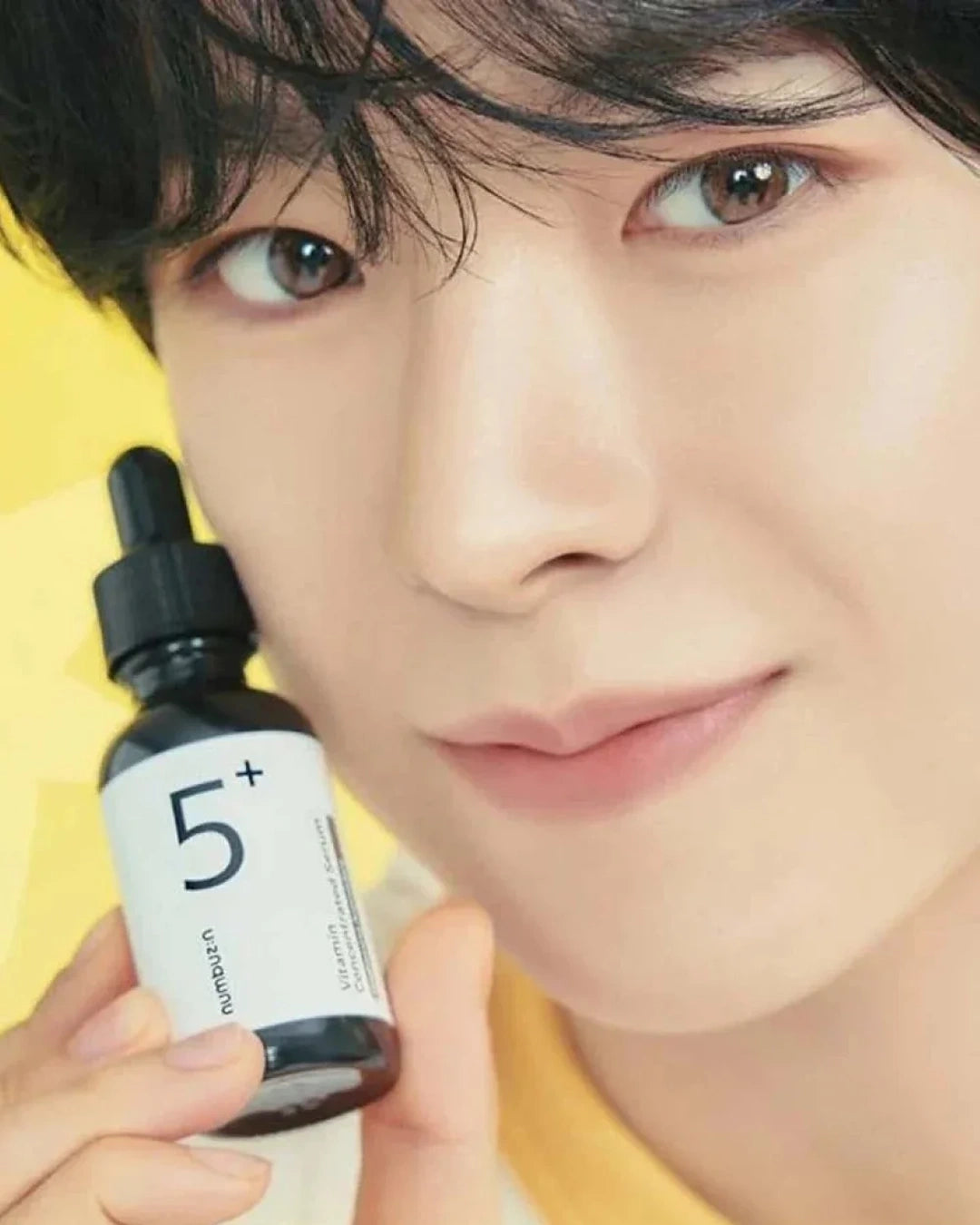 A close-up lifestyle shot of a person holding a small black dropper bottle labeled '5+' against a bright yellow background.