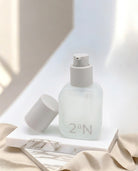 A frosted glass skincare pump bottle with a white cap, shown with its cap off beside it on a white marble block and beige fabric. The bottle has the brand name '2aN' printed on the front.